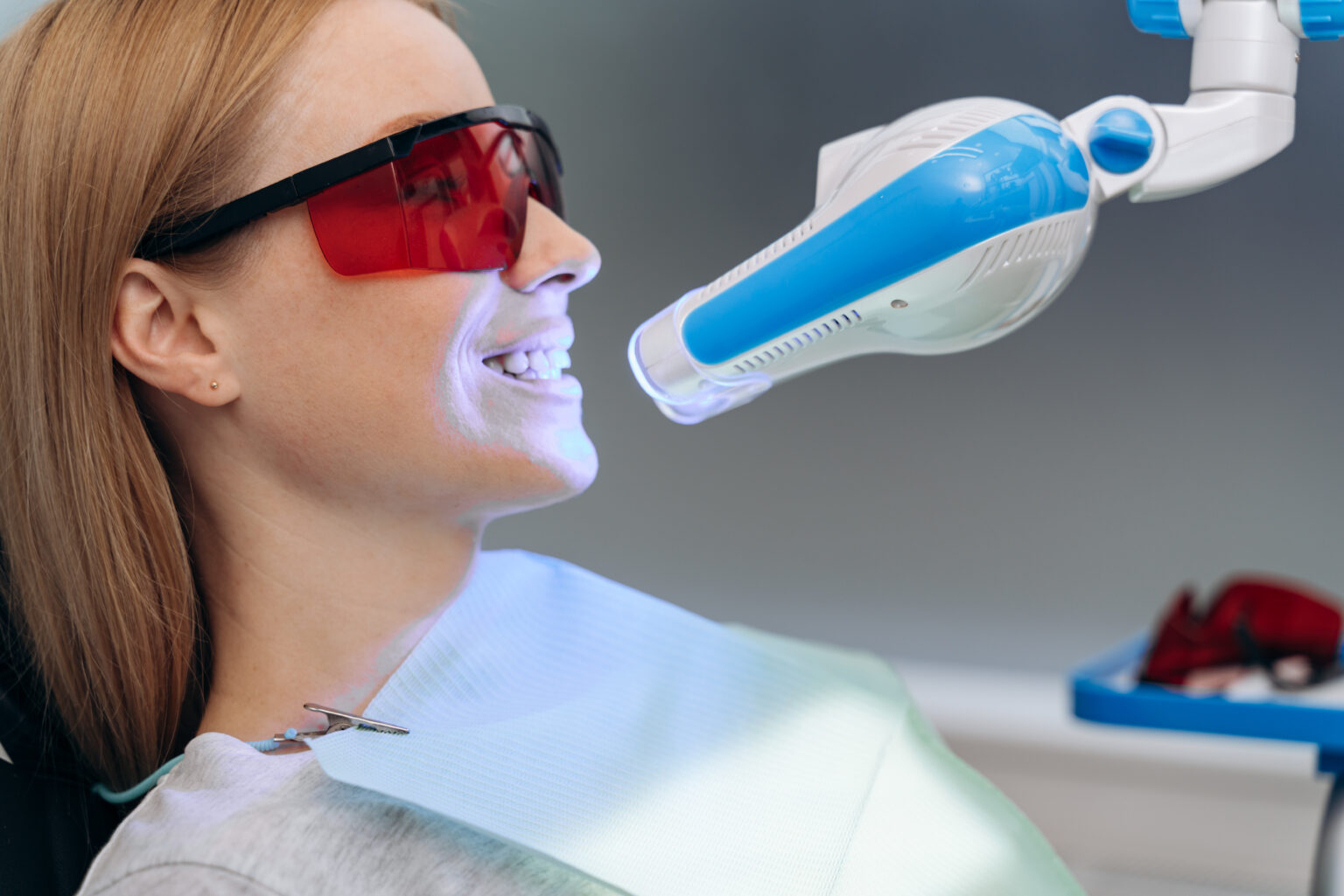 Attractive, young patient in a dental chair with goggles shows her teeth. Dental equipment shines on the patient's teeth